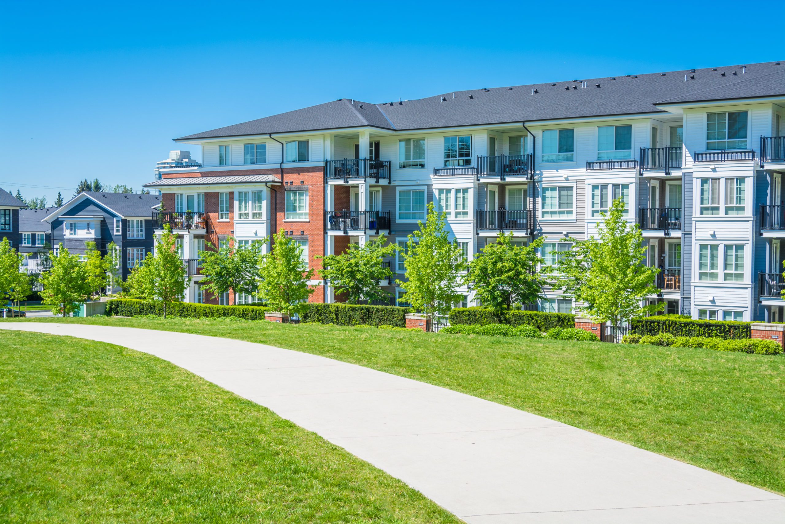 bigstock--152793614 Concrete pathway across green lawn in front of residential condo building. Residential apartment building on sunny day with blue sky.